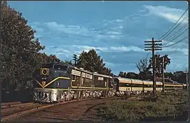 A passenger train with diesel locomotives. The locomotives have blue paint with gold accents and silver undersides. The passenger cars are light gray with blue and gold window stripes.