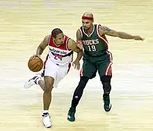 At left man carrying the ball wearing a white Wizards uniform, man at right guarding him wearing a red headband and dark green Bucks uniform