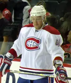 An ice hockey player standing in front of the camera. He is wearing a white helmet and a red, blue and white uniform.