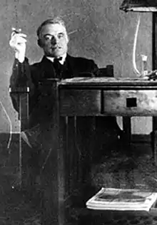 A gray-haired man dressed in a dark suit and white shirt, is looking directly at the photographer. He is sitting in a chair at a table while apparently holding up a cigar.