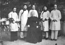 A group portrait of six women, taken outdoors; five Chinese women stand in a row, wearing light-colored tunics and trousers; one white woman, older, is seated in front of the group, wearing all black, with her grey hair dressed in an updo.