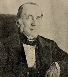 Image of light-skinned man, aged around 65, wearing dark suit and cravat of Victorian period