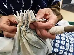 A portion of white fabric with colour-gradient blocks of woven blue pinstripes being tied into cones in the kumo shibori (spider shibori) style by a craftsperson.