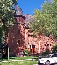 An orange stone building with a rounded tower in front and large pointy-arched entrance in the middle, partially obscured by trees on either side. In the front right a white car is parked in the street.