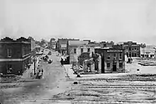 About a dozen one- and two-story buildings, several of which are damaged, line a dirt road that intersects with three railroad tracks in the foreground