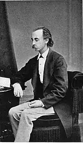 A middle aged man with a mustache, dressed in a tuxedo, seated at a desk