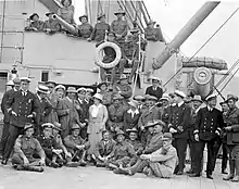 Black and white photo of a group of men and a single woman posing on the deck of a warship. Most of the men are wearing military uniforms.