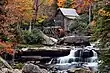 A wooden grist mill standing alongside a rushing stream with a footbridge in the background.