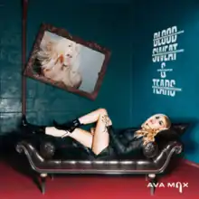 Max is seen laying on a black couch inside a blue room. The song title is written on the right-side of the wall in white paint.