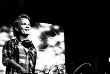 A man standing at a mixing desk on stage smiles as he performs, wearing a striped shirt and a pair of headphones around his neck.