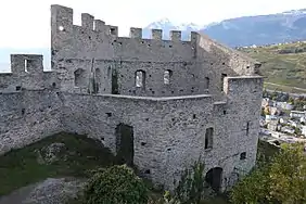 A building without a roof and whose doors are protected by bars. The top of the walls are crenellated.