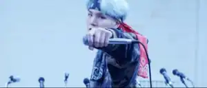 Frontal shot of Suga wearing a dark blue and red bandana around his forehead, looking directly into the camera with his left arm outstretched, holding a silver microphone horizontally in his hand just before he drops it