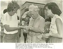 An older white woman with white hair, center, greets a younger white woman, left, while a teenaged girl with dark hair and glasses looks on, right.