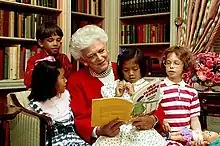 This is a photo of Former First Lady, Barbara Bush, on a couch in the White House Library. She is surrounded by a handful of children crowing her while she reads a book.