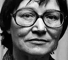 Dark-haired, strong-featured woman in her 30s or 40s, wearing 1970s spectacles.