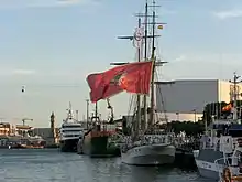 A colour photograph of a sailing ship alongside a dock flying a red flag