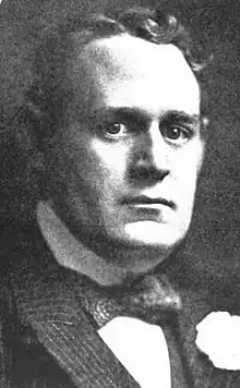 A black-and-white portrait of a Caucasian man looking at the camera, dressed in a striped suit and bow tie.