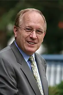 A bespectacled Barry Stuppler in a tan suit, blue shirt, and gold tie.