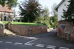 Road junction with house on the right of the road. On the left is a churchyard behind a wall accessed via steps.