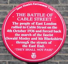 Red plaque from Tower Hamlets Environment Trust reading The Battle of Cable Street. The people of East London rallied to Cable Street on the 4th October 1936 and forced back the march of the fascist Oswald Mosley and his Blackshirts through the streets of the East End. 'They Shall Not Pass'