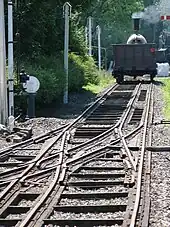 A rail track recedes into the distance where a steam train stands; the track has three rails, the middle of which is offset to the right in the foreground but switches to the left in the middle at some complex pointwork where three other rails join from the left