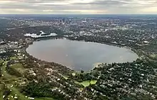 An aerial view of Bde Maka Ska, a mostly round lake with two nubby "legs" at its bottom end, surrounded mostly by parks and houses. Downtown Minneapolis is visible at the top of the image.