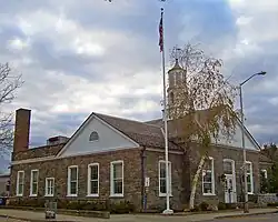 A one-story stone building with white pointed roofs and a cupola in the center underneath a clouded sky. There are a bare tree and a flagpole in front.