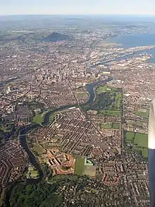 Aerial photo of urban sprawl, edged by green hills and sea shore, and bisected by a winding river.