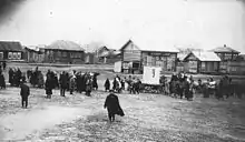 Group of people in a clearing, with wooden buildings in the background