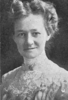 A smiling middle-aged white woman, light hair in a wavy bouffant updo, wearing a lace blouse or dress with a high collar, and a brooch pinned to the throat