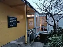 The entrance of the Bill Manhire House, a seemingly small yellow-walled building with stairs down to a lower level and vegetation off to the side.