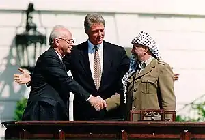 A stolid balding man in a dark suit on the left shakes the hand of a smiling man in traditional Arab headdress on the right. A taller, younger man stands with open arms in the center behind them.