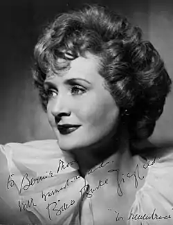 A black and white photograph of a woman with light-colored hair looking off to the left and smiling slightly. It has an ink dedication and signature on the bottom