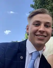 A man with brown hair, wearing a dark blue suit, white shirt and baby blue tie, smiling