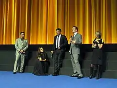 Scott Franklin, Mila Kunis, Vincent Cassel, Darren Aronofsky, and Sandra Hebron stand on a stage with a golden curtain backdrop wearing formal attire and discussing Black Swan