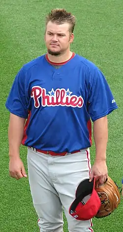 A fair-haired young man wearing a blue baseball jersey and white pinstriped pants stands on a grass field holding a baseball cap and mitt in his left hand