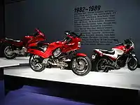 Three sporty motorcycles of the 1990s on pedestals.  Text on the wall behind them says "1982-1989" with a few dozen words relevant to the period, such as "Atari", "MTV", and "Robert Mapplethorpe".