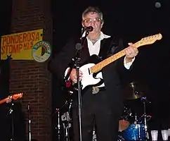 Bob Wootton on stage playing an electric guitar in front of a microphone stand