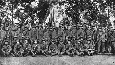 Portrait photograph of a group of men in uniform, posing with rifles and bandoliers