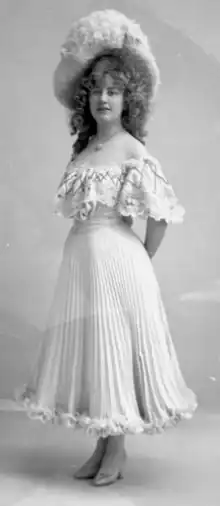 A young white woman, standing, wearing a light-colored pleated and lace-trimmed dress with an off-the-shoulder neckline, and a large hat; her hair is voluminous and falls longer than her shoulders.