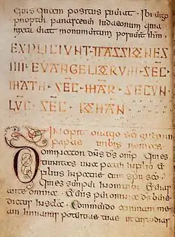 Medieval manuscript leaf bearing 13 lines of Latin script in dark brown & orange ink. Lines 8–13 indent to fit a large initial ‘d’ in dark brown ink with red & green washes. The stem of the ‘d’ transforms into the head of a cat, its long tongue sticking out & tying itself into knots.