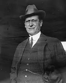 Black and white photo of a white man in a three-piece suit with glasses and a hat.