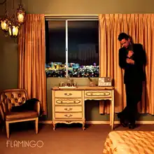 Brandon Flowers stands next to a desk in a hotel room. The windows show the Las Vegas skyline at night.