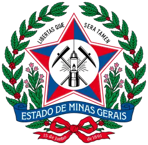 A coat of arms with a large red star in the middle bearing mining equipment. Above the star are the words "Libertas Quae Sera Tamen." The coat of arms is surrounded by a wreath of red berries and green leaves, with two stems of leaves sprouting from the bottom and framing a banner stating "Estado de Minas Gerais." A green ribbon is at the bottom; on the left tail, it says "15 de junho" and on the right, "de 1891."