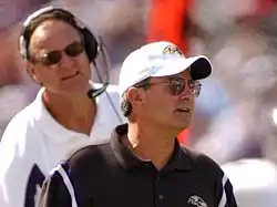 Candid photograph of Zauner standing in front of Brian Billick on a football sideline wearing a black polo shirt and white baseball cap both of which bear the Baltimore Ravens logo