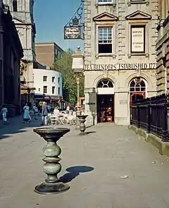 Corn Street, Bristol. A stone-paved pedestrianised street surrounded by classical architecture. In the foreground are two ornate cast bronze tables known as a nails.