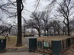 A green fence, about four feet high, opens on a path towards a basketball court, slightly up a hill. In the midground and background, atop lawns of brownish grass stand leafless trees against an overcast sky.