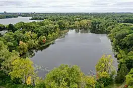 An aerial shot of a small, slightly oblong lake surrounded on all sides by green trees. On the upper lefthand side are some houses and another lake. The sky is overcast.