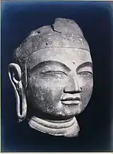 A Buddha head in three-quarter view with elongated earlobe, thick lips, narrow slit eyes and broad nose.