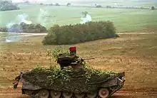 Colour photograph of a tracked military vehicle driving through a rural area